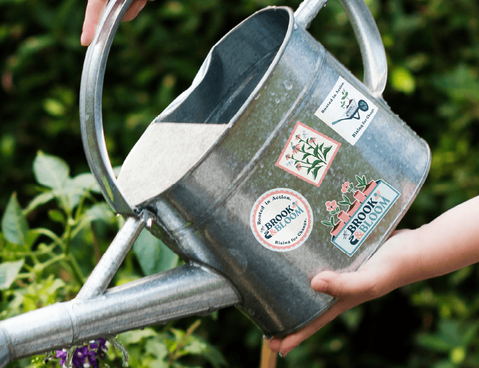 The image depicts a metal watering can with four Brook & Bloom stickers on it.